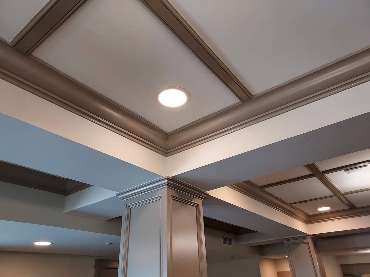 A coffered ceiling with a round recessed light, the beams and crown molding painted a warm taupe against white panels and a square painted column.