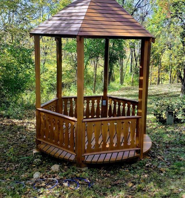 A hexagonal wooden gazebo with a shingled roof and railing, freshly stained a warm amber, standing on a leaf-covered lawn among trees.