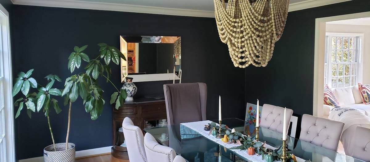 A dining room with deep green walls, white trim, a beaded wood chandelier, a glass-topped table with upholstered chairs and a large leafy plant.